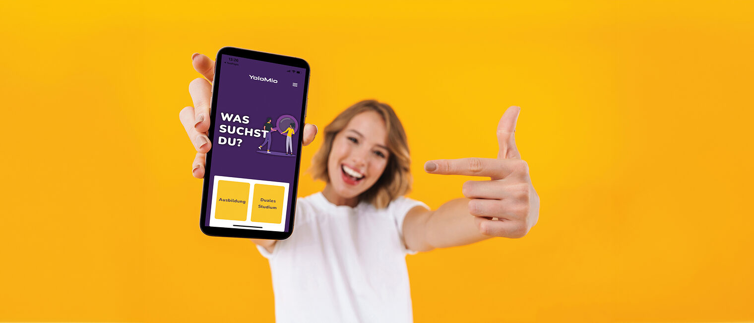 Image of young blond woman in basic t-shirt pointing finger at smartphone in hand isolated over yellow background Schlagwort(e): woman, portrait, smiling, beautiful, excited, joyful, cont