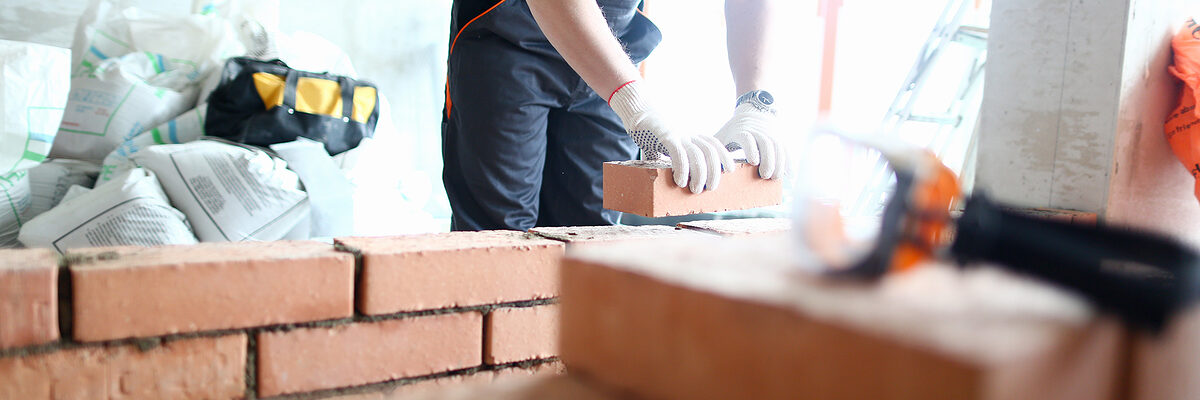 Male builder hand in gloves holding clay brick on background of brickwork wall. Repair and redevelopment apartments concept. Schlagwort(e): industrial, symbol, sign, architecture, object, concept, engineering, technology, line, male, architect, person, service, occupation, renovation, profession, thin, block, structure, foreman, builder, arm, uniform, construction, worker, man, hand, house, repair, home, building, contractor, handyman, constructor, brick, wall, work, build, cement, flat, masonry, brickwork, background, design, bricklayer, professional, workman, industry, maintenance, concrete