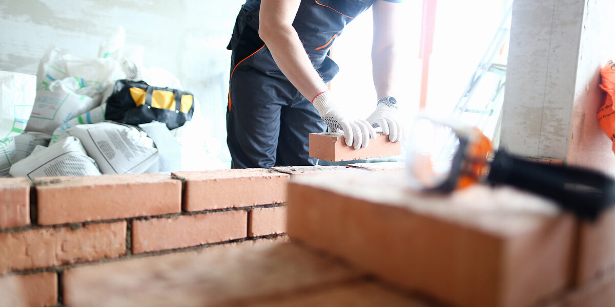 Male builder hand in gloves holding clay brick on background of brickwork wall. Repair and redevelopment apartments concept. Schlagwort(e): industrial, symbol, sign, architecture, object, concept, engineering, technology, line, male, architect, person, service, occupation, renovation, profession, thin, block, structure, foreman, builder, arm, uniform, construction, worker, man, hand, house, repair, home, building, contractor, handyman, constructor, brick, wall, work, build, cement, flat, masonry, brickwork, background, design, bricklayer, professional, workman, industry, maintenance, concrete